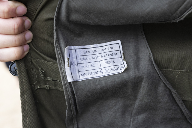 A hand holds open the inner lining of an olive green jacket, revealing a white fabric label with text and care symbols.